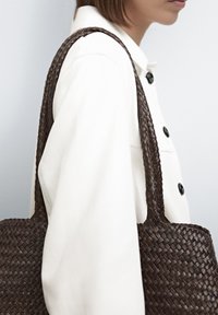 Brown woven tote bag with textured pattern, featuring sturdy braided handles. Worn over a light-colored garment, against a neutral background.
