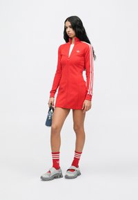 Sieviete stāv sarkana Adidas zip-up kleitā ar baltām svītrām, saskaņotās sarkanās zeķēs, pelēkās sporta kurpēs un turot mazu zilu rokassomu.