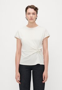 Kortermet kremfarget t-skjorte med en vridd knute-detajl i livet og rund halslinning. Brukt sammen med mørke jeans.