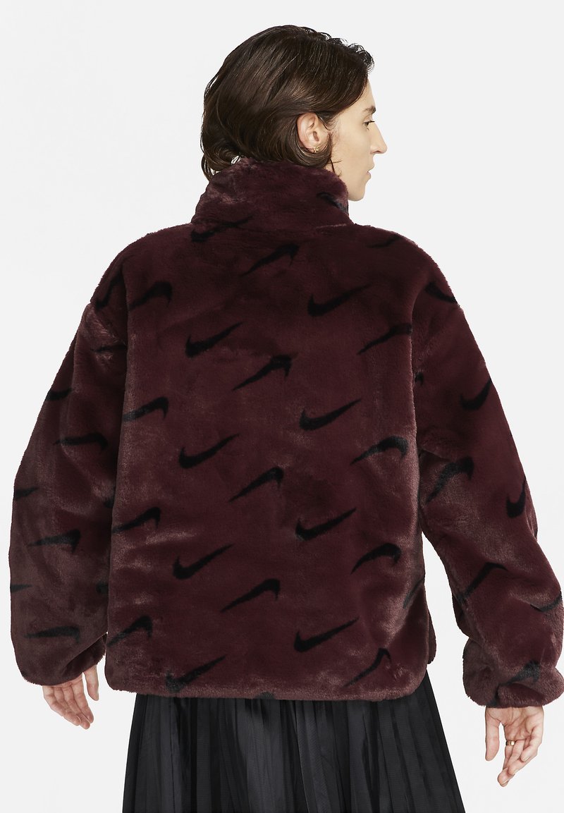 Veste en fausse fourrure bordeaux avec un col haut, une coupe ample et un motif de logo Nike noir sur toute la surface. Texture douce et manches longues.