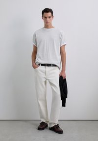 Jonge man in een grijs T-shirt met opgerolde mouwen, witte broek met zwarte riem, die een zwarte trui vasthoudt, staand op een grijze vloer tegen een effen witte muur.