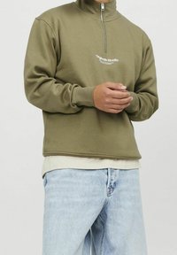 Person som bär en olivgrön tröja med kvartslång dragkedja med texten "Originals Studio", lager på lager över en beige skjorta och ljusblå jeans, med händerna hopknäppta.