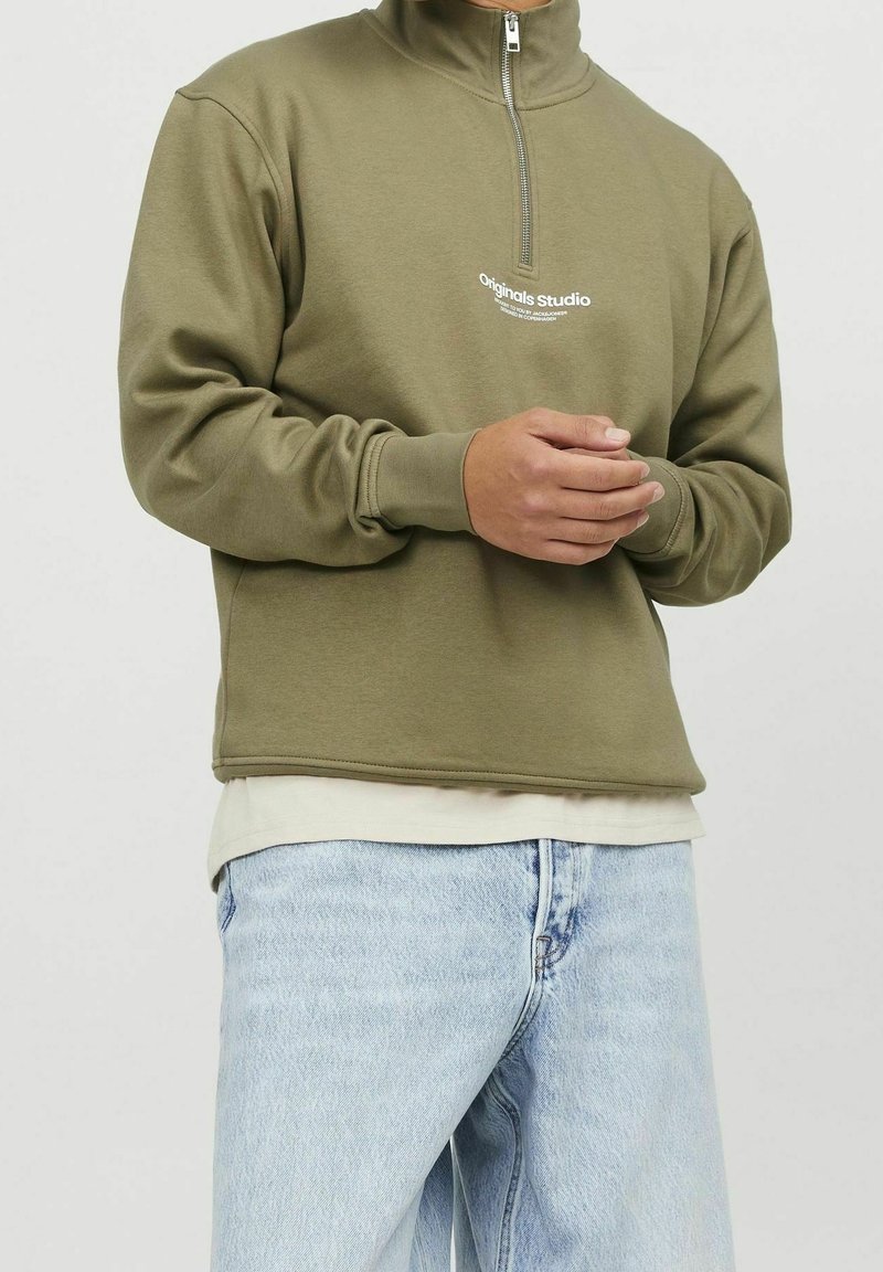 Person som bär en olivgrön tröja med kvartslång dragkedja med texten "Originals Studio", lager på lager över en beige skjorta och ljusblå jeans, med händerna hopknäppta.