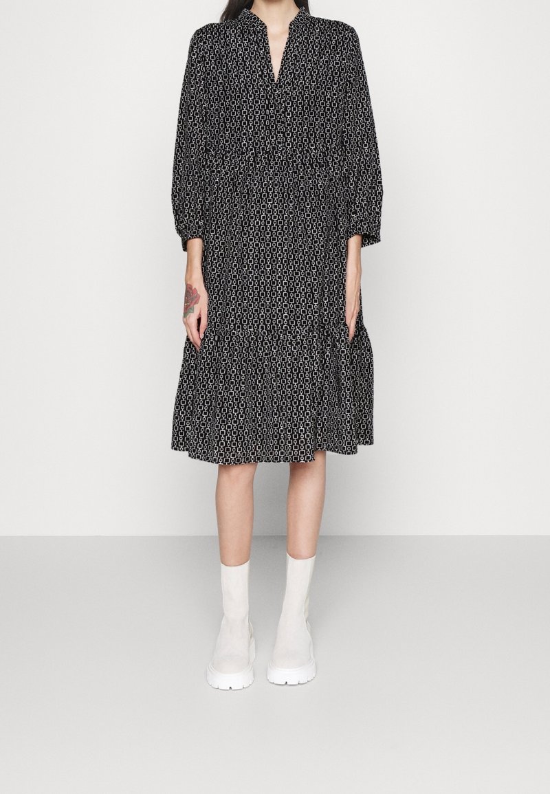Schwarzes und weißes gemustertes Kleid mit V-Ausschnitt, dreiviertel Ärmeln, einem gestuften Saum und einer lockeren Passform. Kombiniert mit weißen Ankle Boots.