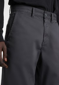 Dunkelgraue Hose mit glatter Textur, ausgestattet mit einem Knopf- und Reißverschluss, seitlichen Taschen und einem taillierten Bund. Minimalistischer Nähdetails.