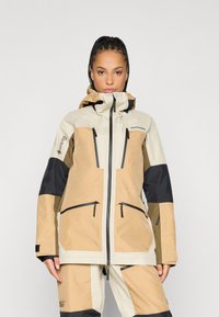 Beige und schwarze Gore-Tex-Jacke mit Kapuze, ausgestattet mit Reißverschlusstaschen und einer taillierten Passform. Kontrastierende Farbblöcke erhöhen das visuelle Interesse.