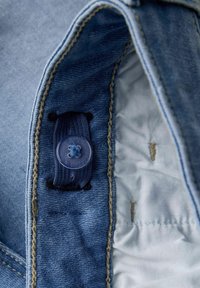 Interno di una giacca di denim con un bottone blu, un elastico navy e cuciture beige su una fodera azzurro chiaro testurizzata.