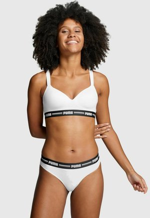 Smiling woman with curly hair wearing white Puma branded bra and matching underwear against plain gray background.