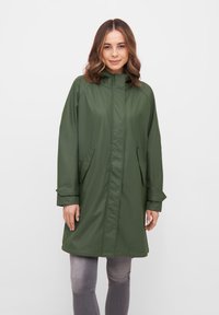 Cappotto impermeabile verde con colletto alto, tasche laterali e maniche lunghe. Texture liscia e taglio dritto, abbinato a jeans grigi.