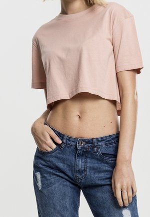 Femme portant un t-shirt court rose et un jean bleu taille haute avec des détails usés, main dans la poche, sur un fond uni.