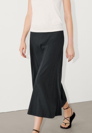 Femme portant un pantalon noir large court, un haut blanc sans manches et des sandales plates noires à lanières marchant sur un sol gris.