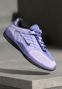 Scarpa sportiva viola con materiale testurizzato, accenti in rete, perforazioni, suola nera piatta e lacci grigi, che presenta un logo argento sul lato.