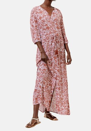 Kobieta ubrana w długą sukienkę z wzorem w odcieniach czerwono-brązowym i białym, z rękawami do połowy przedramienia i beżowymi sandałami, stojąca na jednolitym tle.