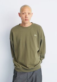 Fiatal kopasz férfi olíva zöld Nike pulóverben és szürke nadrágban, egy sima fehér háttér előtt, kezét a zsebébe dugva.