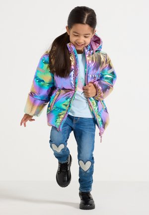 Pige smilende og gående iført en skinnende multicolor puffjakke, lysebluse, jeans med hjertepåstræk og sorte støvler.