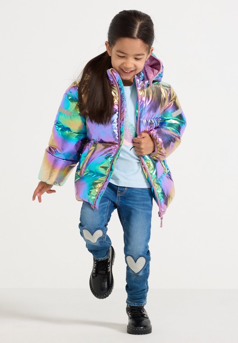 Fille souriante marchant dans une doudoune multicolore brillante, une chemise bleu clair, un jean avec des patches en forme de coeur et des bottes noires.