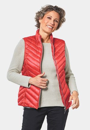 Rote gesteppten Weste mit hohem Kragen und vertikalem Zickzackmuster, mit Reißverschluss auf der Vorderseite, getragen über einem hellgrauen strukturierten Pullover, kombiniert mit dunklen Hosen.