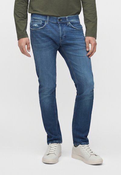 Homme portant un jean bleu slim avec légère usure et des baskets blanches, debout devant un fond uni.