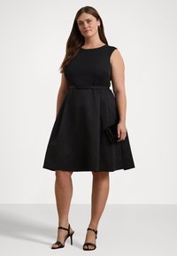 Svart ärmlös klänning med en figurnära överdel, utsvängd kjol, midjebälte och subtil textur. Kombinerad med svarta högklackade sandaler och en kuvertväska.