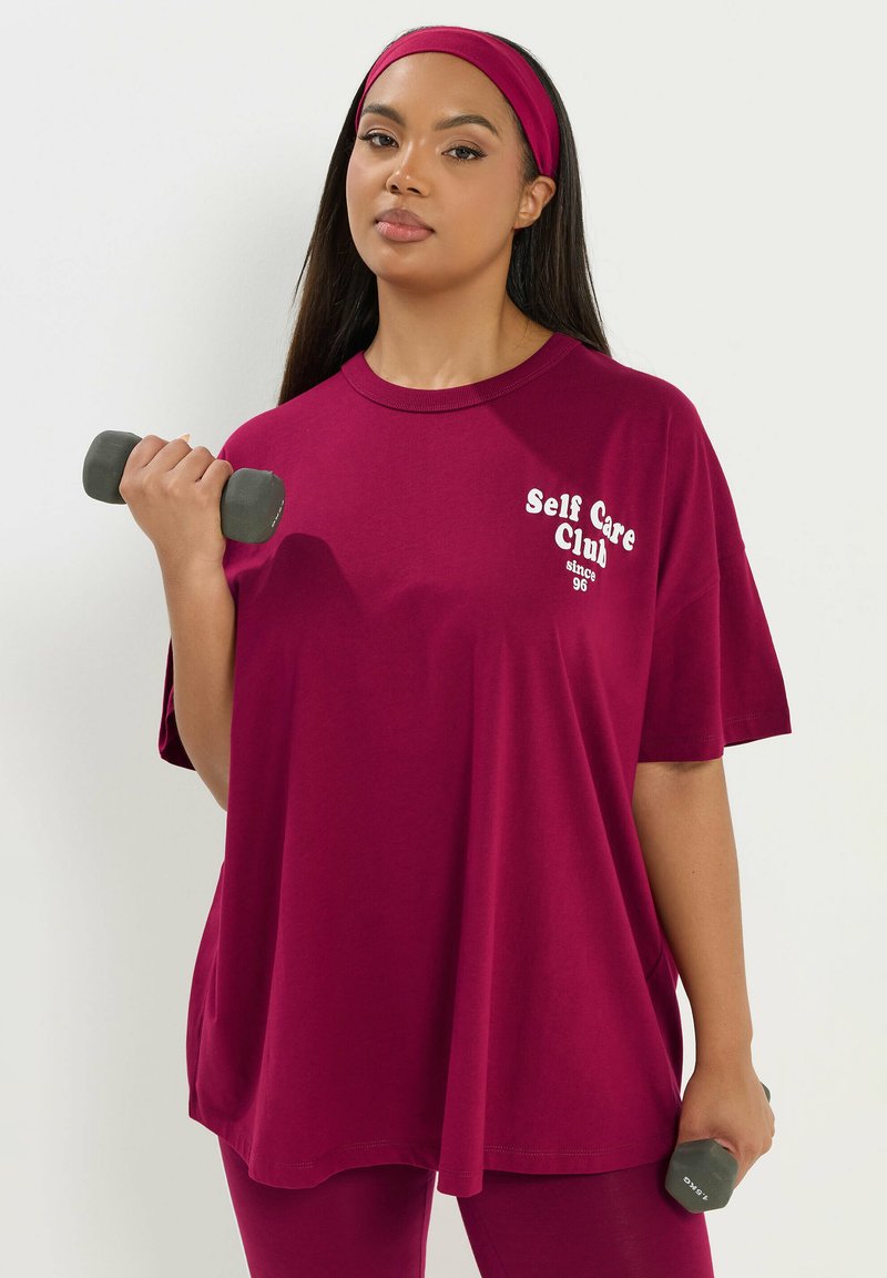 Femme en tenue de sport bordeaux et bandeau tenant deux petits haltères, debout devant un fond clair uni.