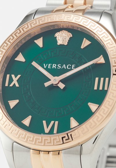 Versace laikrodis su žaliu ciferblatu, auksiniais akcentais ir raštu aplink rėmelį. Yra ryškūs valandų žymekliai ir liūto emblema 12 valandą.