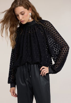 Jonge vrouw met lang golvend haar, die een zwarte getextureerde doorschijnende blouse met ballonmouwen en zwarte leren broek draagt, kijkt naar beneden.