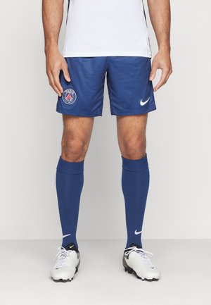 Męski sportowiec w niebieskich spodenkach piłkarskich Paris Saint-Germain, dopasowanych skarpetach, białych korkach i białej koszulce, stoi na jednolitym tle.