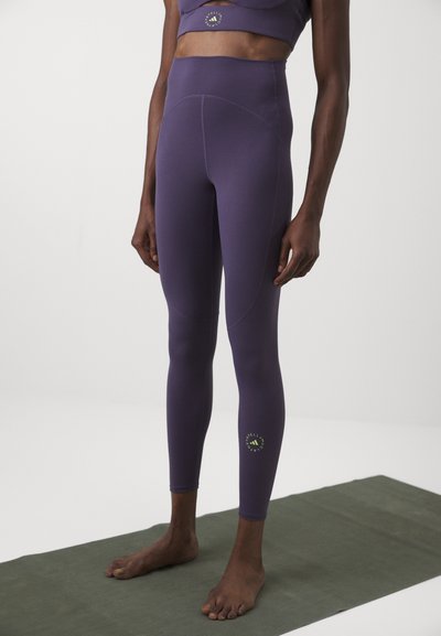 Lila jóga leggings, magas derékkal, sima anyagból készült, ívelt varrásokkal és egy kis zöld logóval a bokánál.