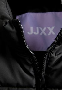 Juodas pūkinis švarkas su užtrauktuku priekyje ir purpurine pamušalu, kuriame užrašyta "JJXX". Medžiaga atrodo lygi ir izoliuota.