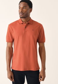 Polo met korte mouwen in gebrande oranje katoen, voorzien van een kraag, drieknopenbies en een klein logo op de linkerborst.