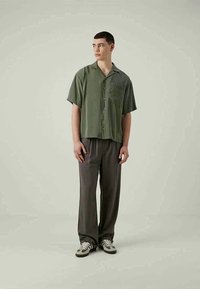 Junger Mann mit kurzen Haaren trägt ein olivgrünes, kurzärmliges, knöpfbares Hemd, dunkle, lockere Hosen und graue Sneakers und steht vor einem schlichten Hintergrund.