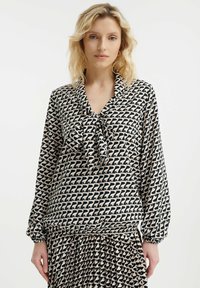 Langmouw blouse in zwarte en crèmekleurige stof met een geometrisch patroon, een strikhalssluiting, en gathered manchetten met geplooide details aan de onderzijde.