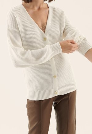 Femme ajustant la manche d’un cardigan en maille blanc à boutons, portant un pantalon marron, un top en dentelle beige visible en dessous.