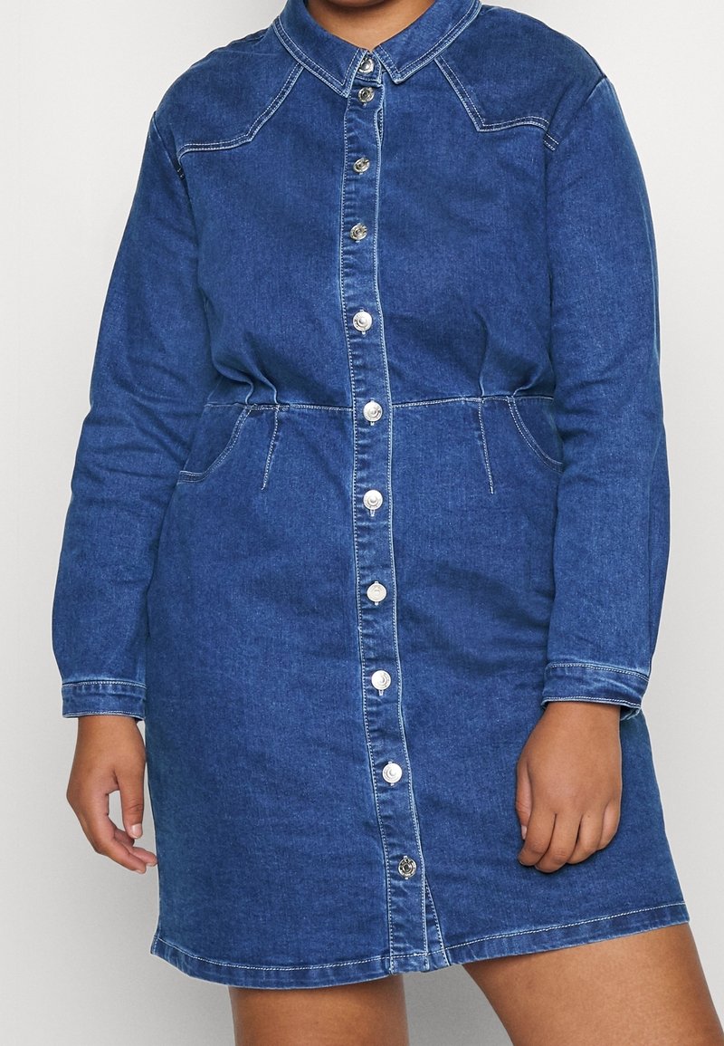 Femme portant une robe en jean bleue à manches longues, boutonnée sur le devant, avec des poches avant et des surpiqûres contrastantes.