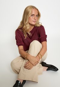 Bordeaux kortemouwen trui met een textuurafwerking, gecombineerd met een beige broek en zwarte instappers. Gouden accessoires accentueren de outfit.