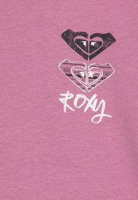 Lyserød bomuldsstof med tre hjerte designs i sort og hvid, med "ROXY" trykt i en håndskrevet stil.
