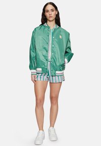 Groene satijnen bomberjack met gestreepte boorden, gedragen over een verticaal gestreept shirt en bijpassende shorts. Witte sneakers maken de look compleet.