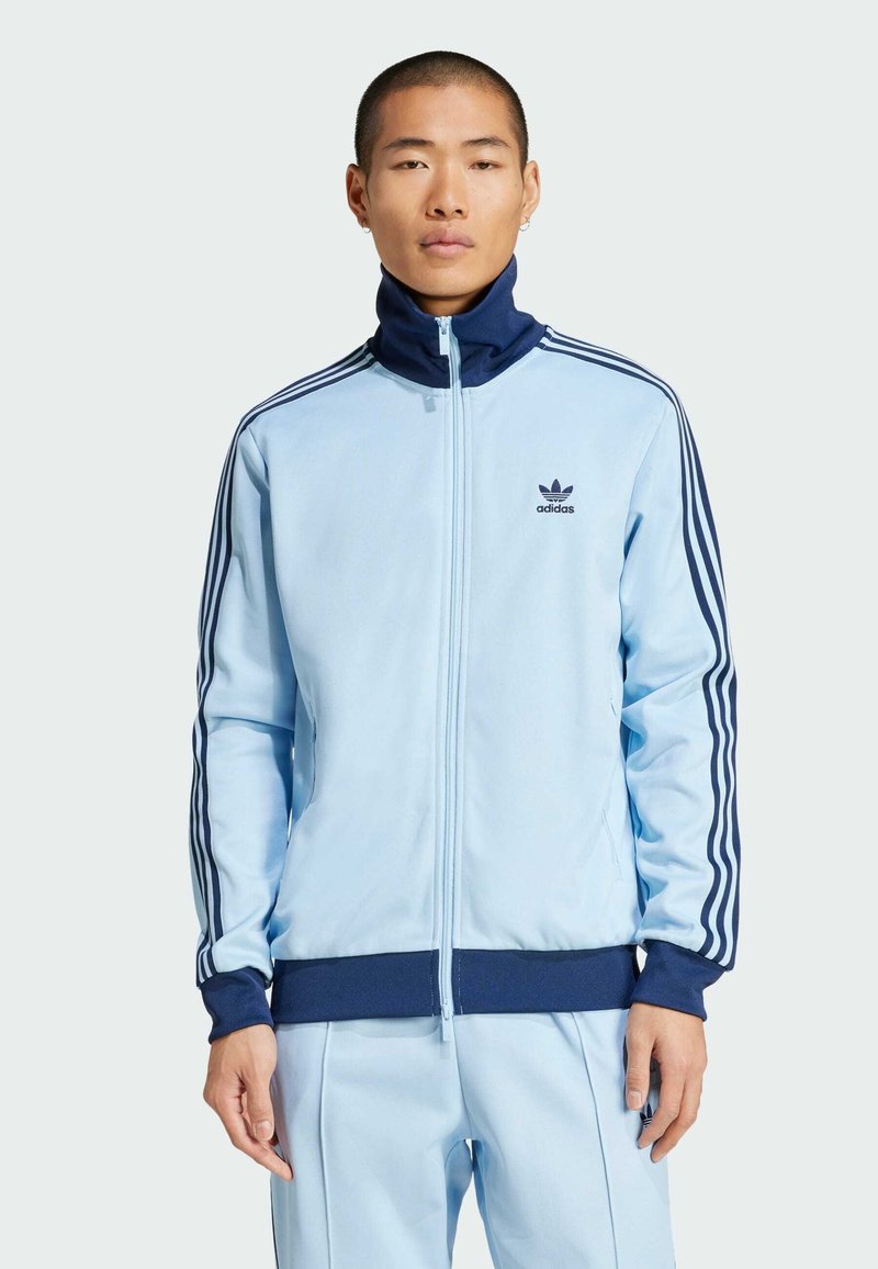 Svijetloplava jakna na zatvaranje s patentnim zatvaračem izrađena od glatkog materijala. Ima tamnoplave detalje i tri pruge duž rukava. Adidas logo na prsima.