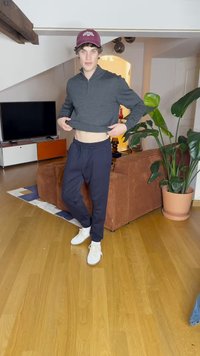 Sweatshirt cinza com fecho, calças azul-marinho com cordão e ténis brancos. Ambiente interior com um sofá de veludo cotelê castanho e uma planta verde num vaso.