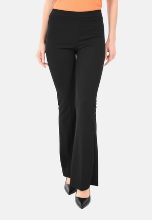 Donna che indossa pantaloni neri a zampa, scarpe nere a punta e un top senza maniche arancione su uno sfondo bianco uniforme.