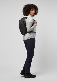 Schwarzer Rucksack mit mehreren Fächern, trägt eine hellgraue, durchsichtige Jacke und dunkle Hosen, steht vor einem neutralen Hintergrund.
