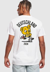 Camiseta blanca con un oso amarillo de dibujos animados jugando al fútbol. El texto en la parte trasera dice "DEUTSCHLAND 2024" en letras negras en negrita, con acentos rojos.