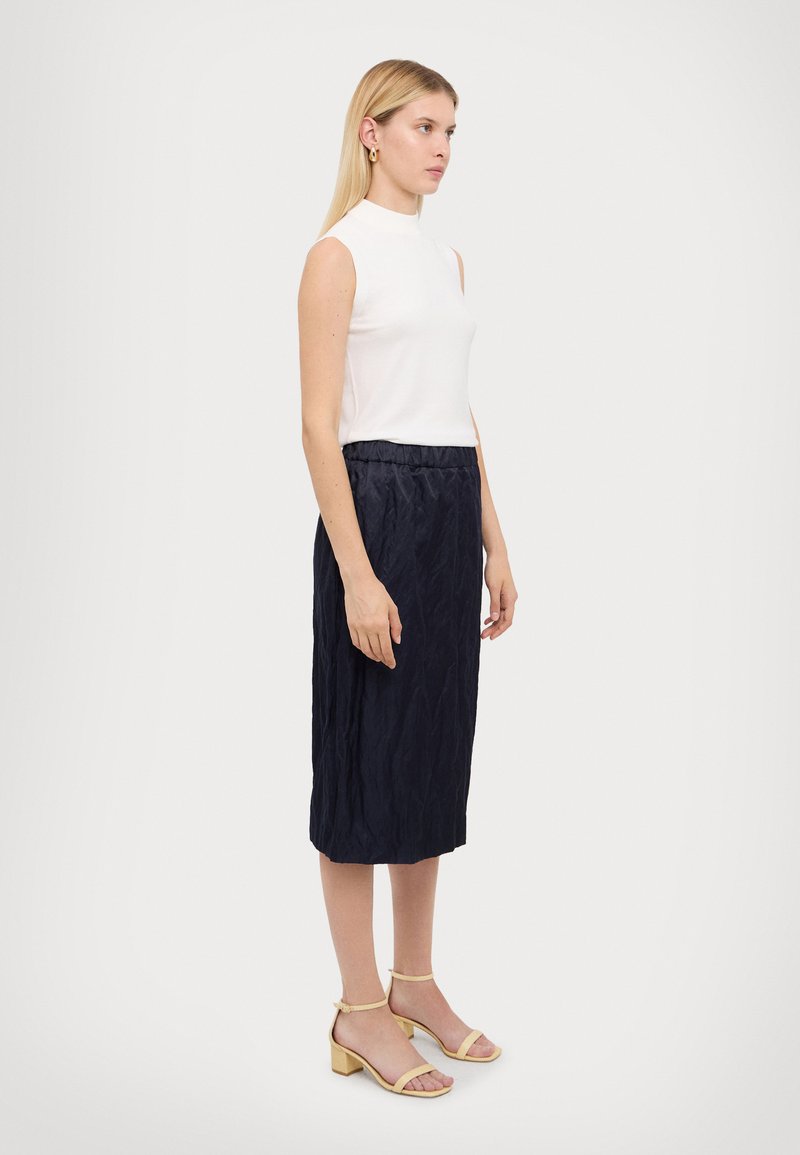 White sleeveless top paired with a navy crinkled skirt. Model wears light yellow block-heeled sandals. Simple, tailored design.