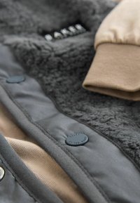 Šedá vnější tkanina s měkkým fleecovým interiérem, béžový náplet a tmavé knoflíky; obsahuje texturovaný vzor, který ukazuje vrstvy a stehy.