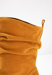 Botte en daim marron au design décontracté, avec un haut arrondi et une petite étiquette en boucle. La texture douce et la finition lisse sont mises en avant.
