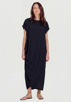 Femme aux longs cheveux foncés portant une robe maxi noire ample et des sandales plates beiges à brides, debout devant un fond clair uni.