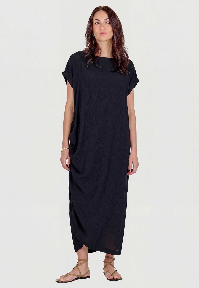 Femme aux longs cheveux foncés portant une robe maxi noire ample et des sandales plates beiges à brides, debout devant un fond clair uni.