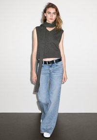 Grijze mouwloze top met een V-hals, gecombineerd met lichtblauwe wijde jeans en witte sneakers, op een model tegen een effen achtergrond.