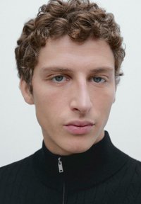 Maglione nero con zip e colletto a coste, caratterizzato da un motivo testurizzato. Primo piano di una persona con capelli ricci chiari e occhi azzurri.