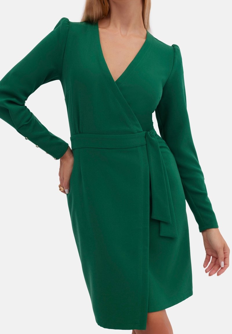 Robe enveloppante verte avec des manches longues, un décolleté en V profond, une taille nouée et des accents de boutons sur les poignets. Tissu lisse avec une coupe structurée.